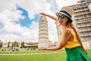 Smiling woman in a straw hat pretends to hold the Leaning Tower of Pisa, a famous tourist attraction in Pisa, Italy.