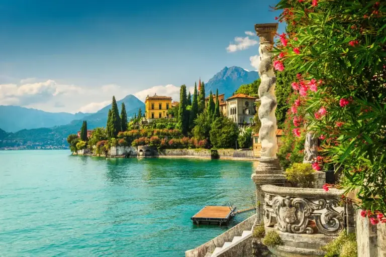 Blooming oleander flowers and elegant sculptures along the lakefront with Villa Monastero in the background on a sunny day at Lake Como, Italy.