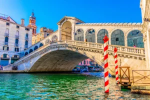 Sunlit view of the iconic Rialto Bridge over the Grand Canal in Venice, Italy, featuring historic architecture and bustling boat traffic.