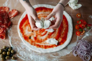 Hands preparing an authentic Italian pizza with fresh mozzarella, spicy salami, and olives on a floured surface, showcasing traditional cuisine in Italy.