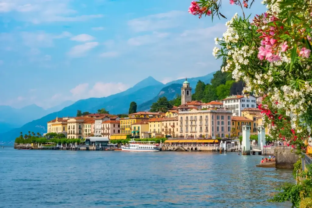 Scenic view of Bellagio, a picturesque town on Lake Como, Italy, showcasing vibrant architecture, a bustling waterfront with a boat, and lush floral foreground.