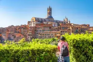 Tourist photographing the panoramic view of Siena including the famous Cathedral Dome and Bell Tower, with lush green foreground, in Tuscany, Italy