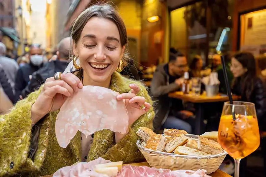 Young woman enjoying traditional cold cuts on a food tour in Bologna, Italy, with a bustling street scene in the background.