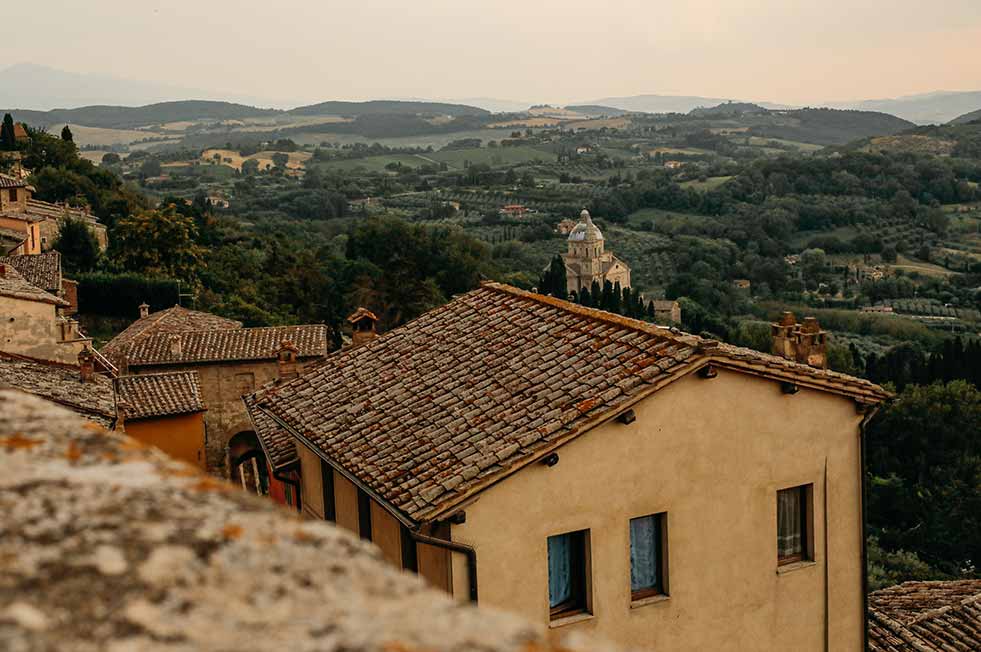 Scenic view of Montepulciano, Italy showcasing historic terracotta rooftops with lush Tuscan hills and a distinctive church in the background, illustrating a quintessential Italian landscape.