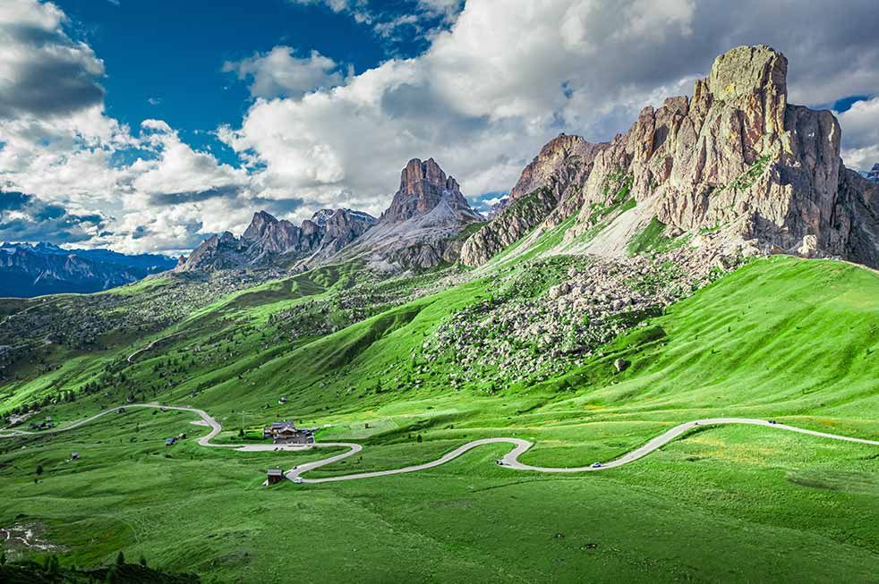 Aerial view of Passo Giau and Averau peak in the lush green Dolomites showcasing winding roads and dramatic rock formations under a dynamic sky.