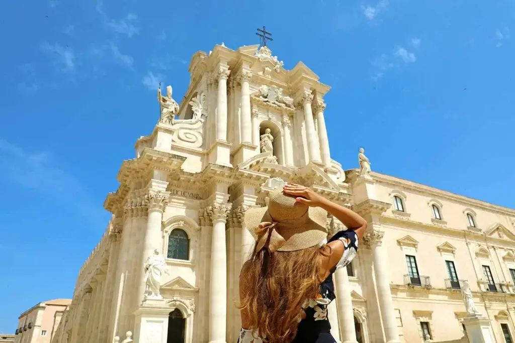 Tourist admiring the intricate baroque architecture of Syracuse Cathedral in Sicily, Italy, under a clear blue sky.