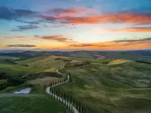 Sunset over the rolling hills of Val d'Orcia, Tuscany, featuring a winding road lined with cypress trees and rich green fields under a vibrant sky.