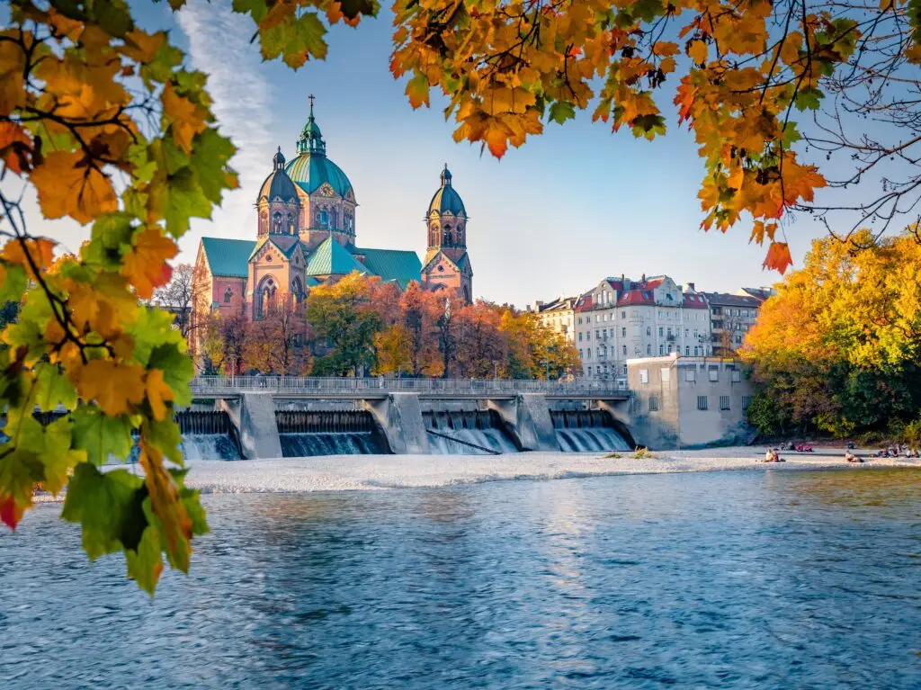 Colorful autumn view of St. Luke's Church in Munich Germany with fall foliage and reflections on the Isar River perfect for Europe private tours