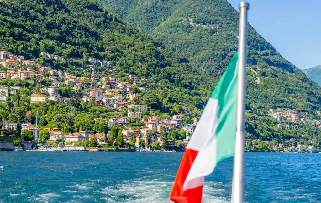 Italian flag waving on a boat cruising Lake Como with colorful hillside villages and lush green mountains in Lombardy, Italy