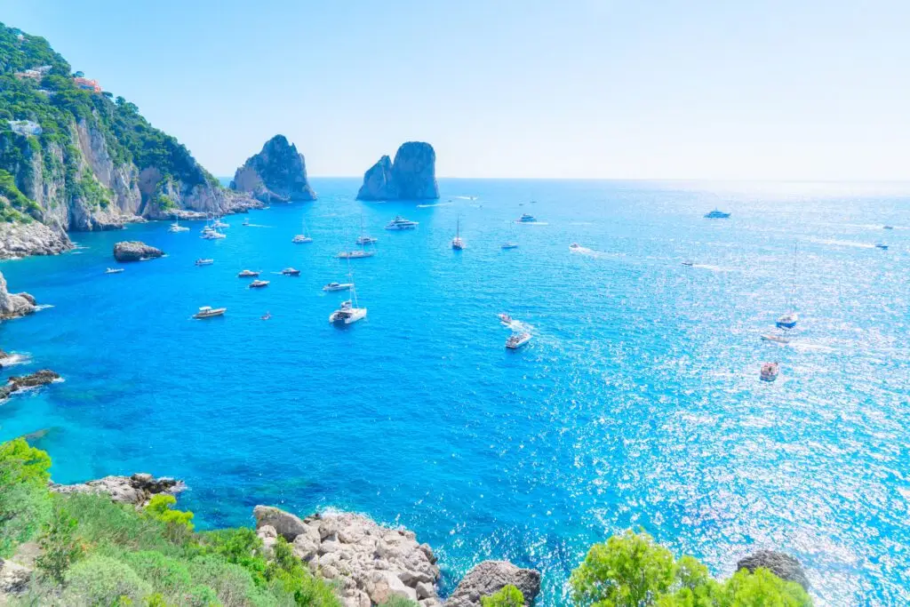 Boats sailing near the Faraglioni cliffs on the Capri Island coast in Italy with turquoise blue sea and rocky shoreline, popular Mediterranean travel destination