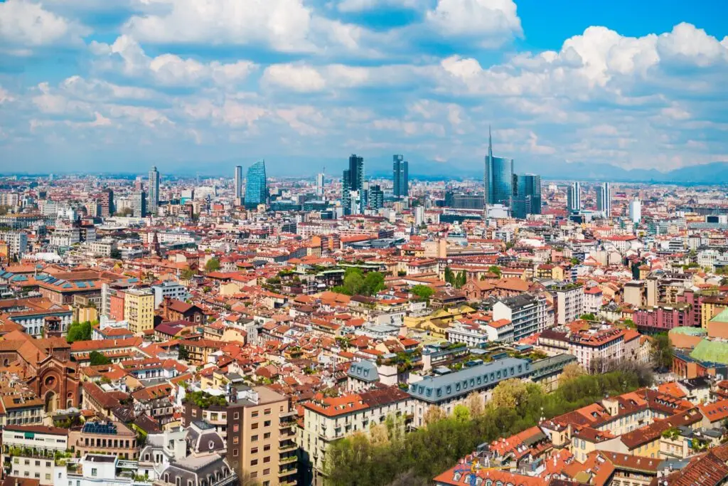 Aerial panoramic view of Milan skyline in Lombardy, Italy, with modern skyscrapers and historic red rooftops under a cloudy blue sky