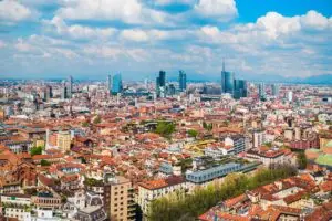 Aerial panoramic view of Milan skyline in Lombardy, Italy, with modern skyscrapers and historic red rooftops under a cloudy blue sky