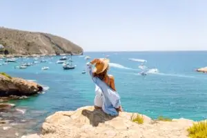 Young woman in sun hat sitting on a cliff overlooking a turquoise Mediterranean bay with boats on an Italy coastal tour